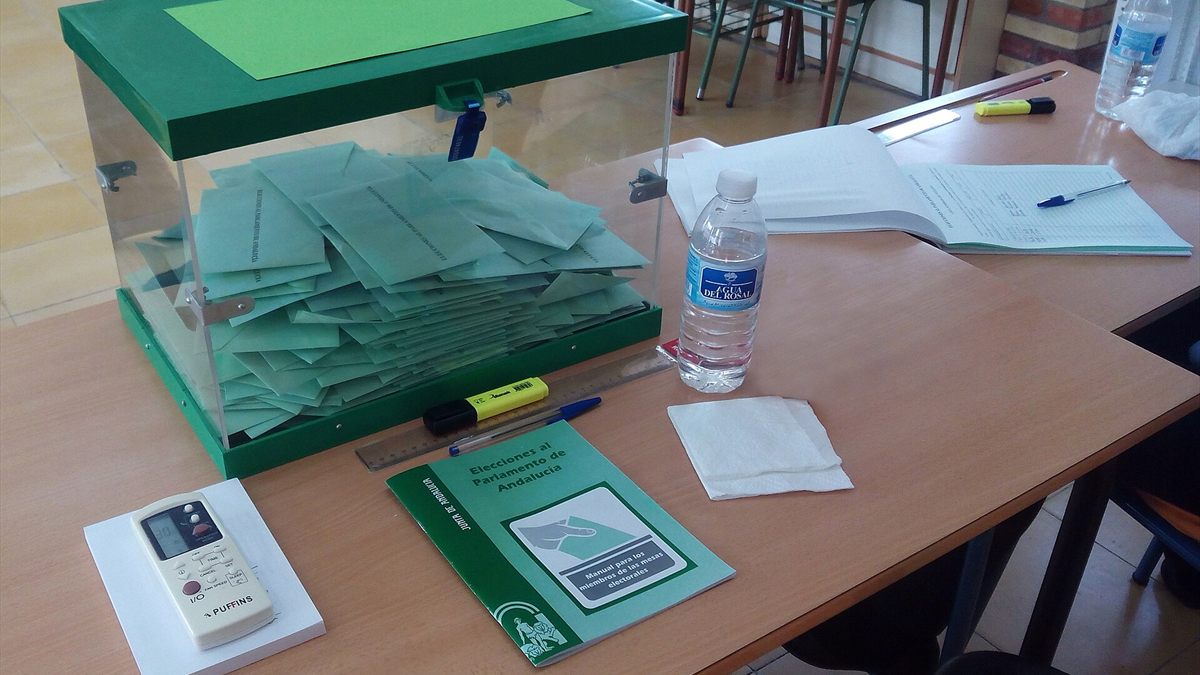 Urna transparente con sobres de voto verde en la mesa de un colegio electoral, junto al manual de la jornada electoral